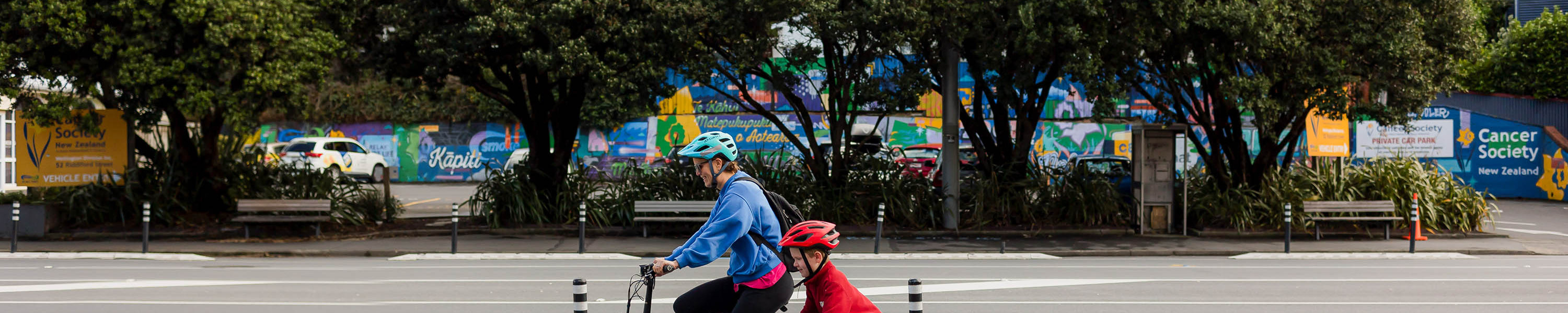An adult and a child ride bikes along a marked bike lane on a Wellington city street, with trees and colourful murals in the background.